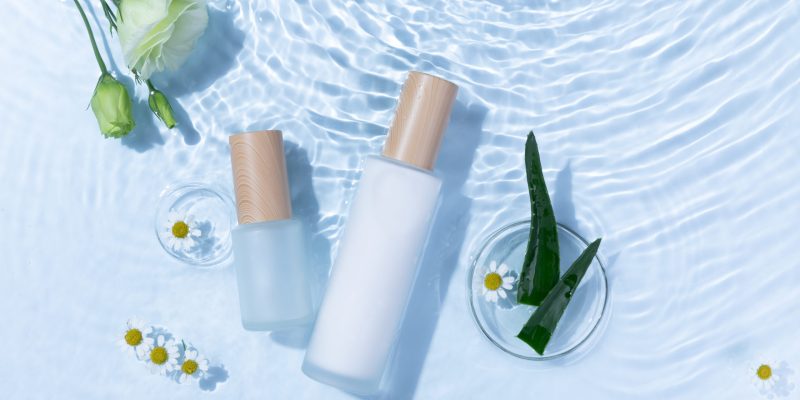 A top view of skincare bottles on a light blue water surface with aloe vera and daisy flowers